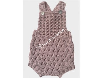 May include: A dusty rose-colored knitted baby romper with a diamond pattern. The romper features a V-neck design with two small buttons on the straps. The text "@punto_bebe" is visible on the garment.