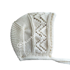 Puede incluir: Gorro de bebé de punto color marfil con un patrón de zigzag decorativo y pequeños detalles de perlas. El gorro tiene un cierre de lazo y el texto "@punto_bebe".