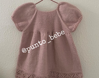 May include: A pink knitted baby dress with short sleeves and a scalloped hem. The dress has a simple design and is perfect for a special occasion. The text "@punto_bebe" is visible on the dress.