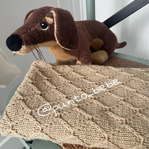 Puede incluir: Un juguete de peluche de perro salchicha marrón y beige descansa sobre una manta de punto beige con un patrón de diamantes. La manta tiene el texto "@punto_bebe" en blanco. El perro tiene una nariz negra y orejas largas.