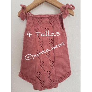Puede incluir: Un pelele de bebé de punto rosa polvoriento con un patrón de diamantes y correas anudadas. El pelele está colgado de una percha de madera. El texto "4 Talllas" y "@punto_bebe" están escritos en el pelele.