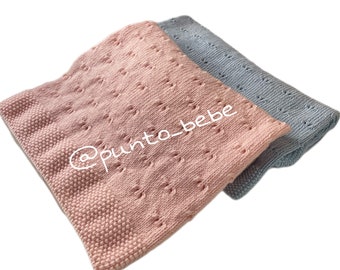 May include: Two knitted baby blankets, one pink and one blue, with a delicate openwork pattern. The blankets are folded together and the pink blanket has the text "@punto_bebe" on it.