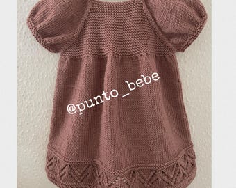 May include: A mauve knitted baby dress with puffed sleeves and a scalloped hem. The dress has a simple design and is made with a soft, comfortable yarn. The text "@punto_bebe" is visible on the dress.