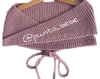 May include: A dusty rose knitted cowl with a tie closure. The cowl is displayed on a wooden hanger. The text "@punto_bebe" is embroidered on the cowl.