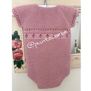 Puede incluir: Un body de bebé de punto rosa empolvado con mangas cortas, colgado de una percha blanca. El body presenta un diseño calado decorativo en el pecho y el texto "@punto_bebe".
