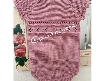 May include: A dusty rose-colored knitted baby romper with short sleeves, hanging on a white hanger. The romper features a decorative openwork design across the chest and the text "@punto_bebe".