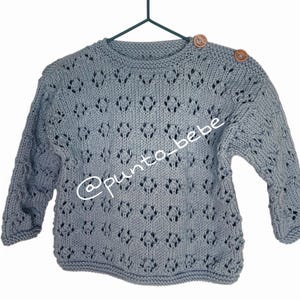Puede incluir: Un suéter de punto azul claro de manga larga con un intrincado patrón de diamantes calados en el cuerpo y las mangas. La prenda incluye dos botones redondos de madera en el hombro izquierdo y el texto @punto_bebe en la parte frontal.