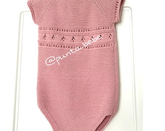 May include: A light pink knitted baby romper with short sleeves. The romper features a decorative openwork design across the chest and the text "@punto_bebe". The garment is hanging on a white hanger.