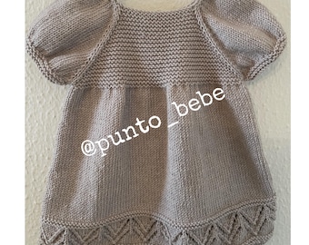 May include: A light brown knitted baby dress with short puffed sleeves and a scalloped hem. The dress has a simple design and is made with a soft, comfortable yarn. The dress is tagged with the social media handle @punto_bebe.