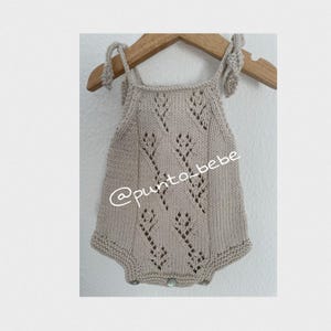 Puede incluir: Un pelele de bebé de punto beige con un delicado patrón de encaje y tirantes de lazo. El pelele tiene una marca de agua @punto_bebe.