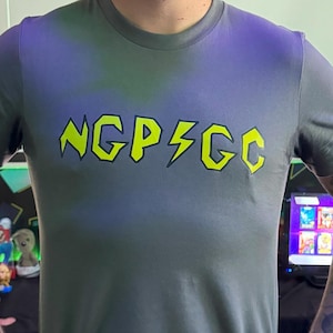 May include: A gray short-sleeve t-shirt with the text "NGP⚡GC" in neon yellow. The person is wearing glasses and jeans. The background includes a projector screen and a wall with geometric shapes.