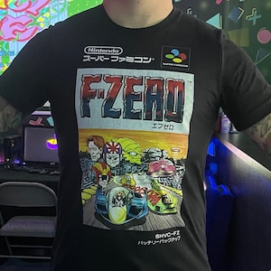 May include: A person wearing a black t-shirt with a graphic of the video game F-Zero on the front. The shirt has the text "Super Famicom" and "F-Zero" in Japanese. The person is standing in a room with a video game arcade in the background.