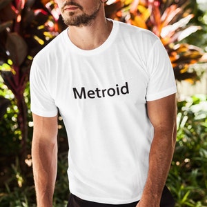 May include: A white t-shirt with the word "Metroid" printed in black on the front. The person wearing the shirt is standing in front of a green leafy background.