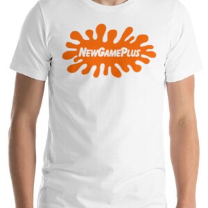 May include: White t-shirt with an orange splatter design and the text "NEW GAME PLUS". The shirt is made of a soft material and is suitable for casual wear. The model is wearing grey trousers.