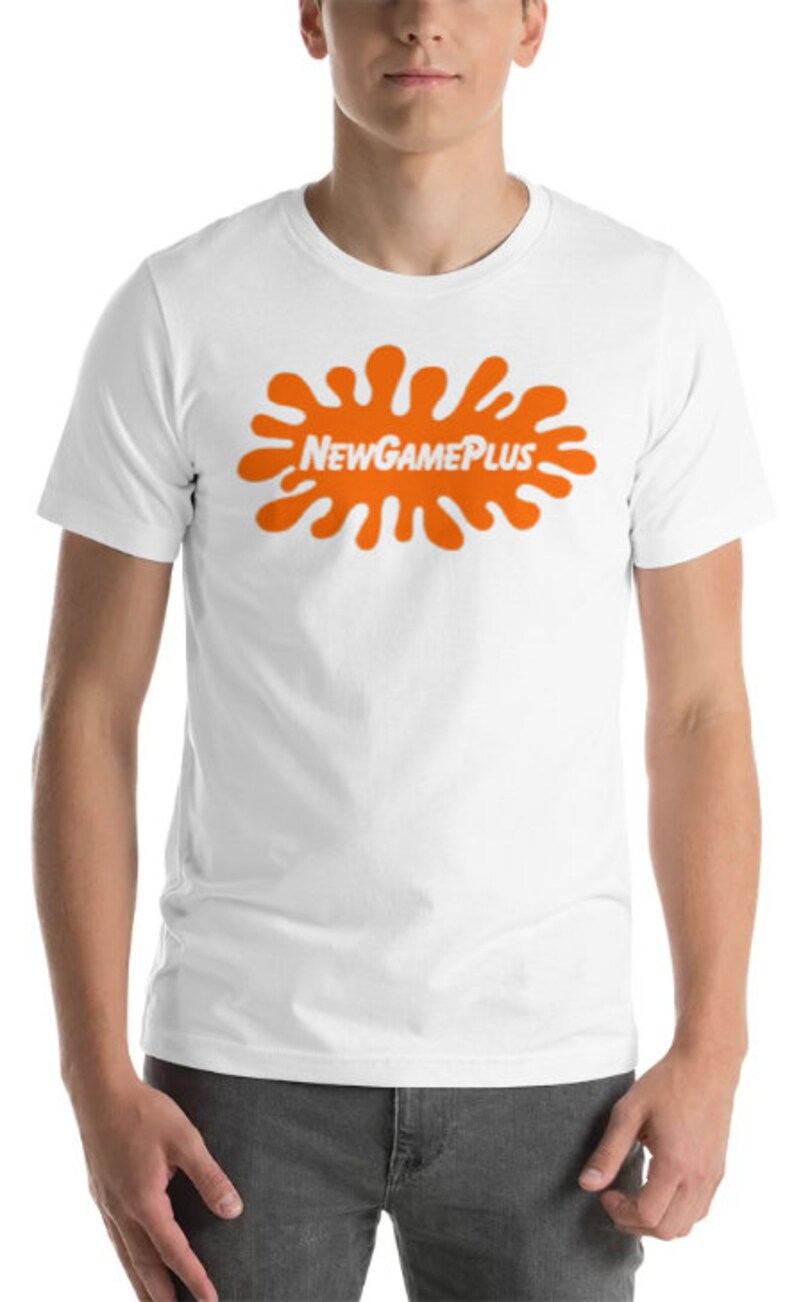 May include: White t-shirt with an orange splatter design and the text "NEW GAME PLUS". The shirt is made of a soft material and is suitable for casual wear. The model is wearing grey trousers.