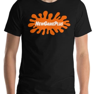 May include: Black t-shirt with an orange splatter design and the white text "NewGamePlus". The short-sleeved shirt has a crew neck. The design is centered on the chest.