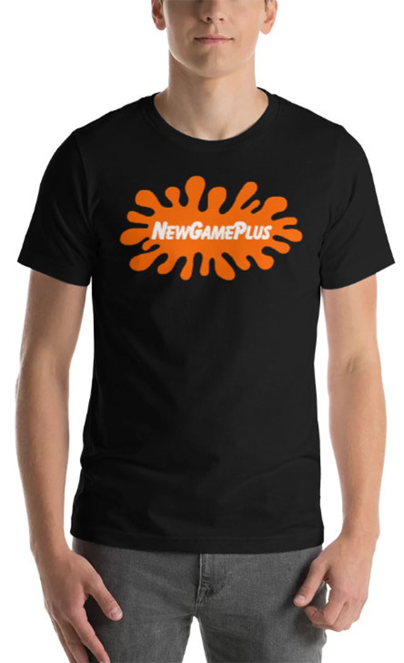 May include: Black t-shirt with an orange splatter design and the white text "NewGamePlus". The short-sleeved shirt has a crew neck. The design is centered on the chest.