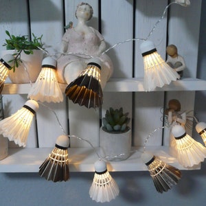 Könnte beinhalten: Dekorative Lichterkette mit Lampen in Form von Federbällen. Die Federbälle wechseln sich mit weißen und braunen Federn ab, mit einem schwarzen Band oben. Die Lichter sind auf einem dünnen Silberdraht aufgereiht und erzeugen ein warmes Leuchten.