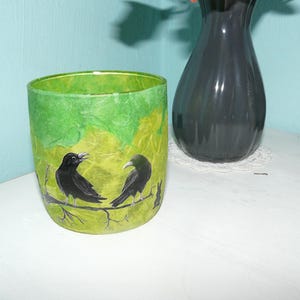 Könnte beinhalten: Ein grüner Glas-Kerzenhalter mit einem bemalten Design von zwei schwarzen Krähen auf einem Ast. Der Hintergrund hat ein strukturiertes, geschichtetes Aussehen mit Grün- und Gelbtönen.