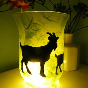 Könnte beinhalten: Ein dekorativer Glas-Kerzenhalter mit einer Silhouette einer Ziege und eines Zickleins. Der Kerzenhalter wird von innen beleuchtet und erzeugt einen warmen, gelben Schein. Das Glas hat ein strukturiertes, grün getöntes Aussehen.
