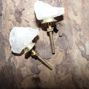 Könnte beinhalten: Zwei Schrankknöpfe mit einer natürlichen, grob geschnittenen Quarz-Kristall-Oberseite. Die Knöpfe haben einen goldfarbenen Sockel und eine Gewindestange zur Montage. Der Hintergrund ist eine strukturierte, braune Oberfläche.