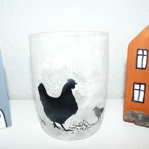 Könnte beinhalten: Ein durchsichtiger Glas-Kerzenhalter mit einer schwarzen Silhouette einer Henne und eines Kükens. Holzhausdekorationen in Blau und Orange umgeben den Kerzenhalter. Der Kerzenhalter ist etwa 13 cm hoch.