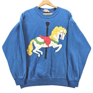 Puede incluir: Sudadera azul con un diseño de caballo de carrusel. El caballo es blanco con crin y cola amarillas, una silla de montar roja y detalles verdes y blancos. La sudadera tiene cuello redondo y mangas largas.