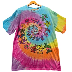 Pode incluir: Uma camiseta tie-dye com um design em espiral em tons de azul, rosa, laranja e amarelo. A camisa apresenta um padrão repetido de ursos dançando em várias cores. A camisa está pendurada em um cabide de madeira.