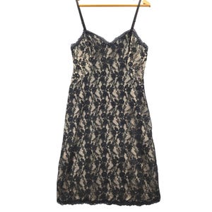 May include: A black lace slip dress with adjustable spaghetti straps. The dress features a scalloped neckline and hem, and is displayed on a wooden hanger. The dress is a neutral color and is made of a delicate lace fabric.