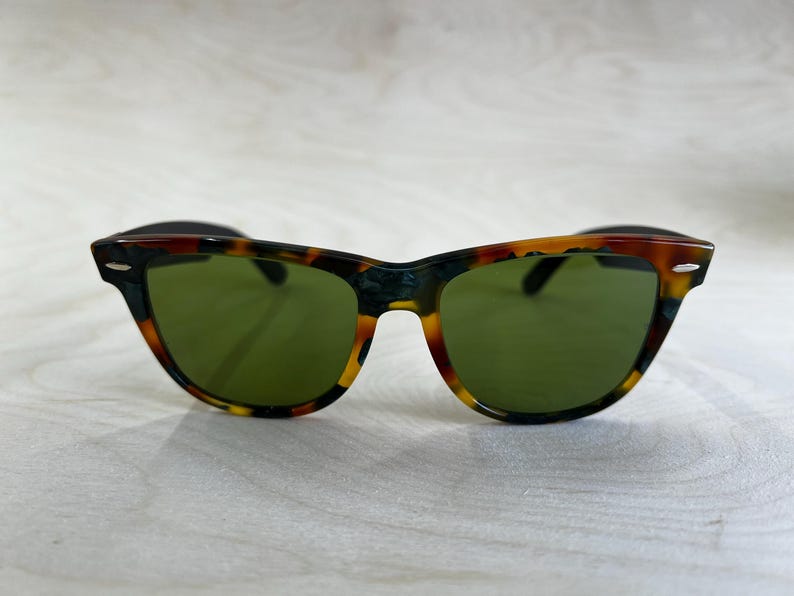 K&ouml;nnte beinhalten: Sonnenbrille mit Schildpattmuster und gr&uuml;nen Gl&auml;sern. Die Fassungen haben ein klassisches Design mit einer Mischung aus Braun-, Schwarz- und Gelbt&ouml;nen. Diese Sonnenbrille ist ein stilvolles Accessoire.