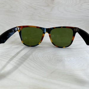 K&ouml;nnte beinhalten: Sonnenbrille mit einem Rahmen im Schildpattmuster und gr&uuml;nen Gl&auml;sern. Die B&uuml;gel sind schwarz. Die Sonnenbrille ist vor einem hellen Holzmaserungshintergrund platziert.