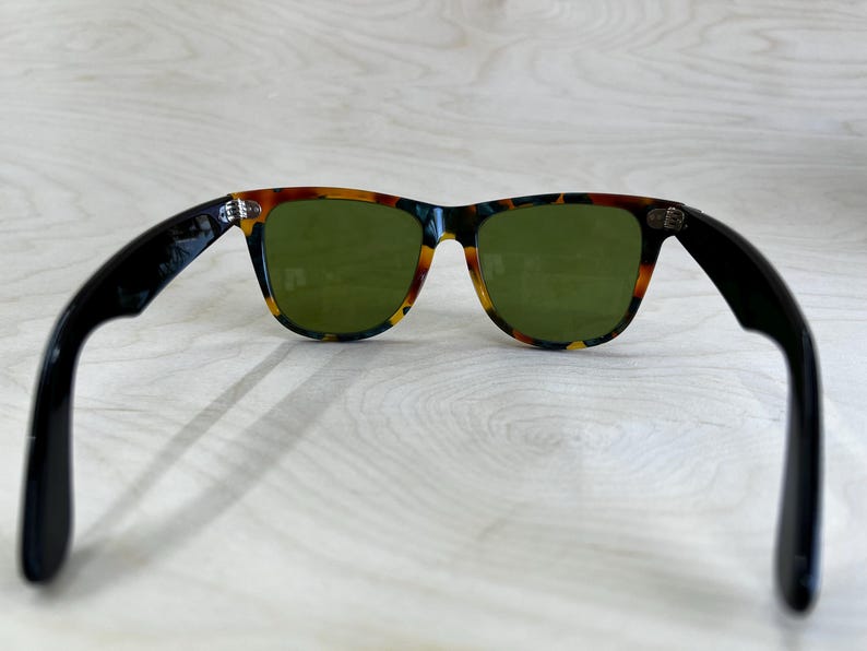 K&ouml;nnte beinhalten: Sonnenbrille mit einem Rahmen im Schildpattmuster und gr&uuml;nen Gl&auml;sern. Die B&uuml;gel sind schwarz. Die Sonnenbrille ist vor einem hellen Holzmaserungshintergrund platziert.