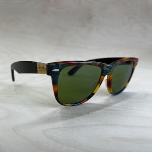 K&ouml;nnte beinhalten: Sonnenbrille mit einem Rahmen im Schildpattmuster und gr&uuml;nen Gl&auml;sern. Die B&uuml;gel sind schwarz mit einem goldenen Logo. Die Sonnenbrille ist vor einem hellen Holzmaserungshintergrund platziert.