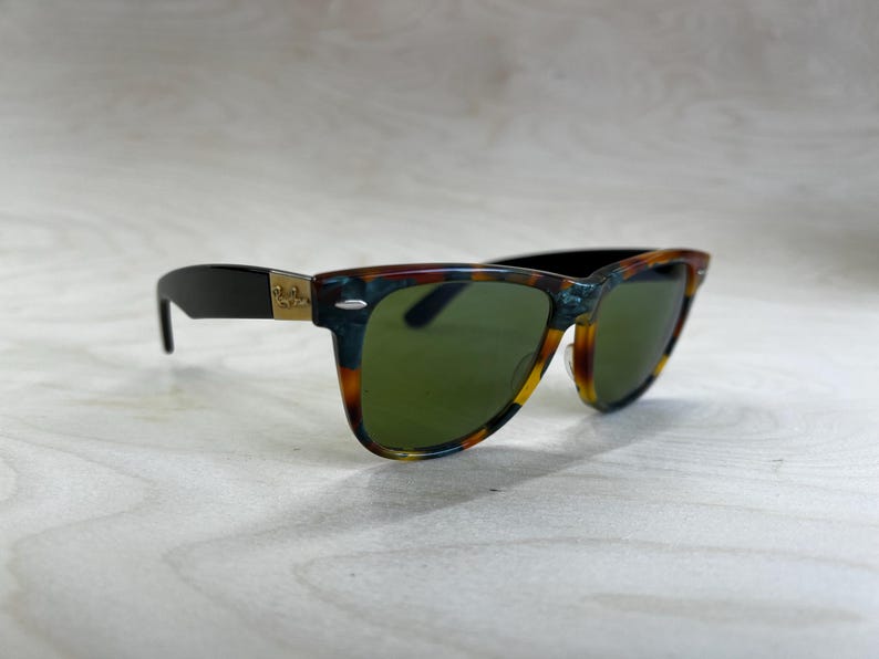 K&ouml;nnte beinhalten: Sonnenbrille mit einem Rahmen im Schildpattmuster und gr&uuml;nen Gl&auml;sern. Die B&uuml;gel sind schwarz mit einem goldenen Logo. Die Sonnenbrille ist vor einem hellen Holzmaserungshintergrund platziert.