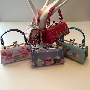 Pode incluir: Seis pequenas carteiras com diferentes desenhos de cupcakes. As carteiras são feitas de um material macio e têm um fecho de metal. As carteiras são de diferentes cores, incluindo rosa, azul, roxo e branco.