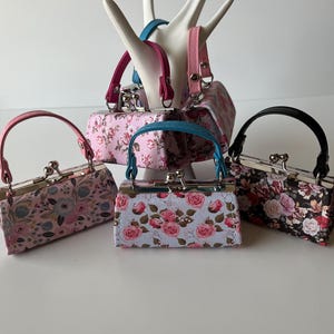 以下が含まれることがあります： 金属製の留め具とハンドルが付いた、花柄の小さな財布が4つ。財布は積み重ねられており、一番上の財布は白い手の形をしたスタンドの上に置かれています。