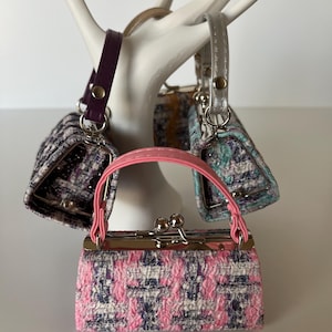 May include: A selection of small handbags in different colours and patterns. The bags are displayed on a white, hand-shaped stand. One bag is pink and white, with a pink handle. Other bags have purple, silver, and brown straps.
