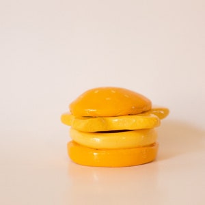 May include: A yellow ceramic hamburger with a sesame seed bun, cheese, and a bottom bun. The hamburger is made of clay and is a decorative item.