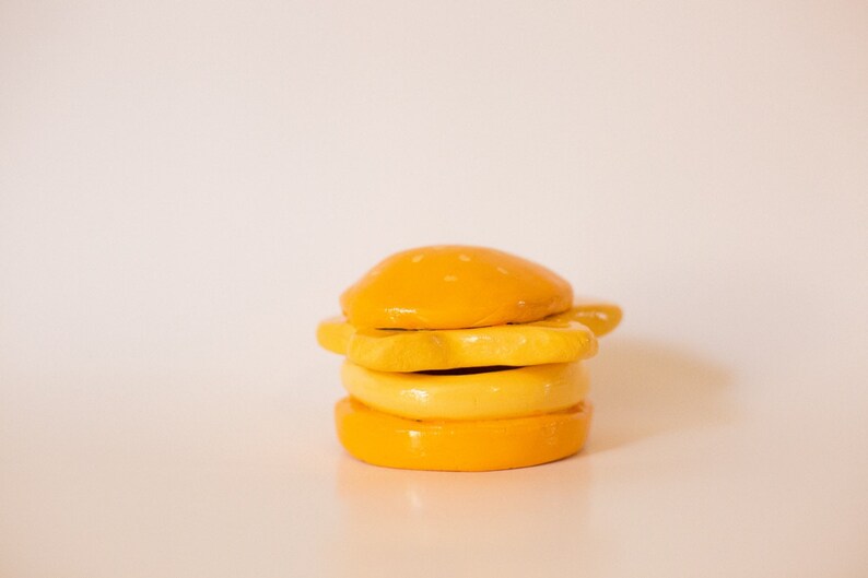 May include: A yellow ceramic hamburger with a sesame seed bun, cheese, and a bottom bun. The hamburger is made of clay and is a decorative item.