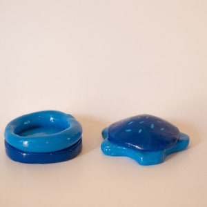 May include: Two blue ceramic trinket boxes. One is a round bowl shape with a slightly raised rim. The other is a turtle shape with a blue and white speckled design.