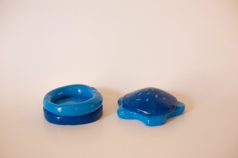 May include: Two blue ceramic trinket boxes. One is a round bowl shape with a slightly raised rim. The other is a turtle shape with a blue and white speckled design.
