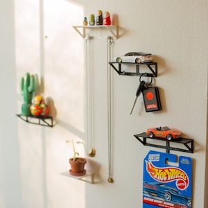 May include: Three black metal shelves mounted on a white wall. The top shelf has a set of nesting dolls, the middle shelf has a white toy car, and the bottom shelf has an orange toy car and a Hot Wheels car packaging. The packaging has the text "Hot Wheels" and "Sugar Rush Series".