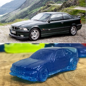 May include: A dark green toy car model of a BMW 3 series, with a black front bumper and black side skirts. The car is sitting on a wooden surface next to a blue toy car model.