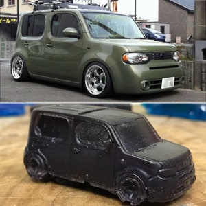 May include: A black toy car model of a Nissan Cube, a small, boxy car. The toy car is sitting on a wooden surface.