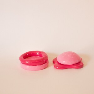 May include: Two pink and red clay sculptures, one is a ring with a hollow centre and the other is a dome shape with a flat base.