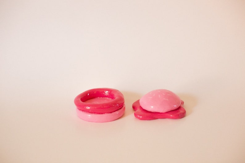 May include: Two pink and red clay sculptures, one is a ring with a hollow centre and the other is a dome shape with a flat base.