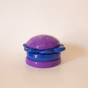 May include: A purple and blue clay sculpture of a hamburger. The top bun is purple and has a star-shaped design. The bottom bun is purple and the patty is blue.