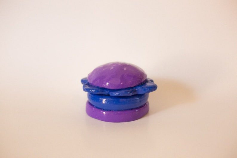 May include: A purple and blue clay sculpture of a hamburger. The top bun is purple and has a star-shaped design. The bottom bun is purple and the patty is blue.