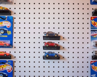 Puede incluir: Un tablero de clavijas blanco con una variedad de coches de juguete Hot Wheels expuestos en estantes. Los coches son rojos, azules y amarillos. El tablero de clavijas tiene un logotipo de Hot Wheels azul y blanco en el lado derecho.