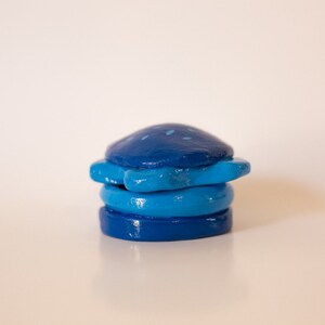 May include: A stack of four blue and teal ceramic coasters with a starburst design. The coasters are painted with a speckled pattern.